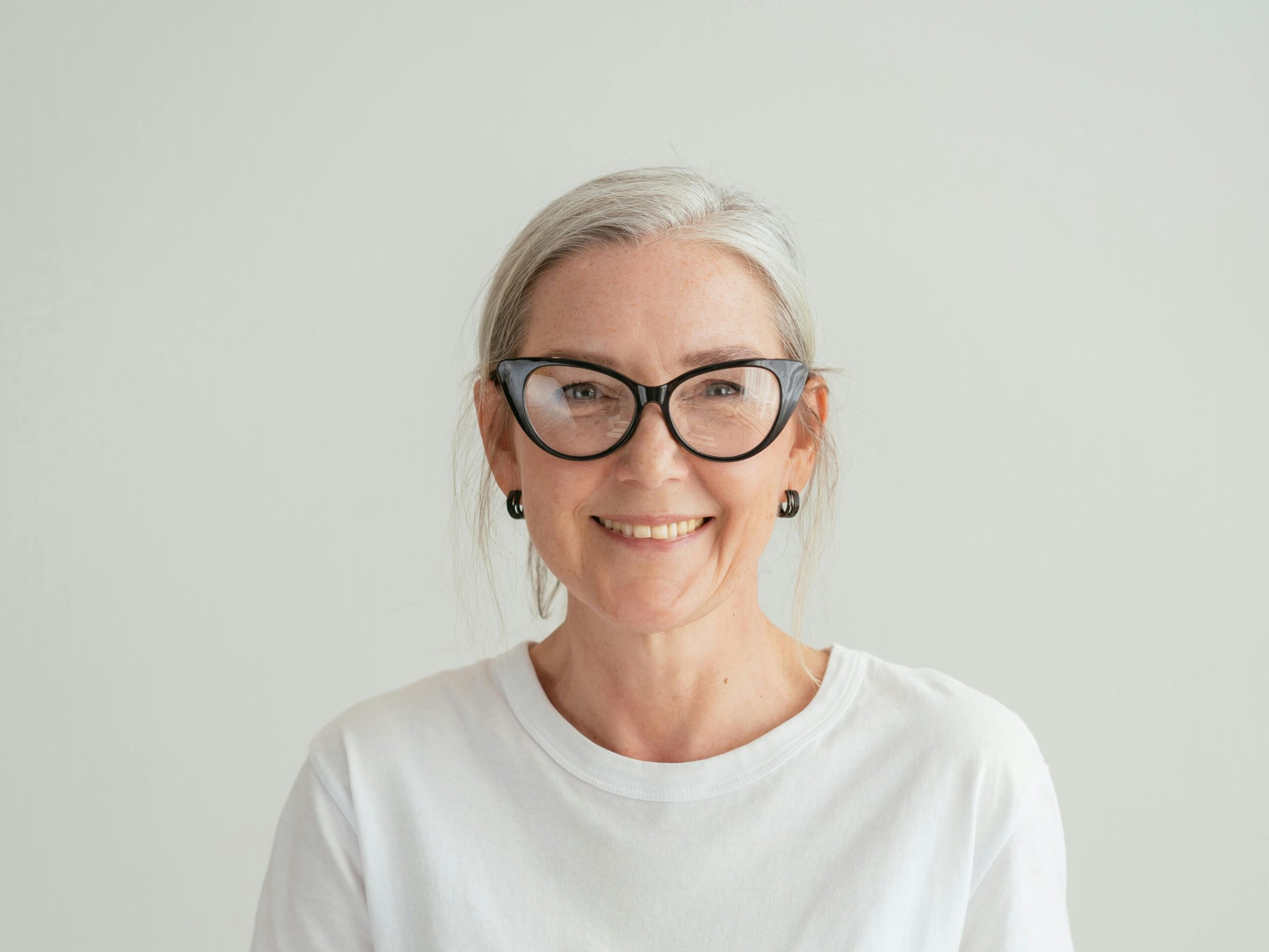 A smiling older woman with gray hair, black-framed glasses, and a white t-shirt stands against a plain light-colored background. A smiling older woman with gray hair, black-framed glasses, and a white t-shirt stands against a plain light-colored background.