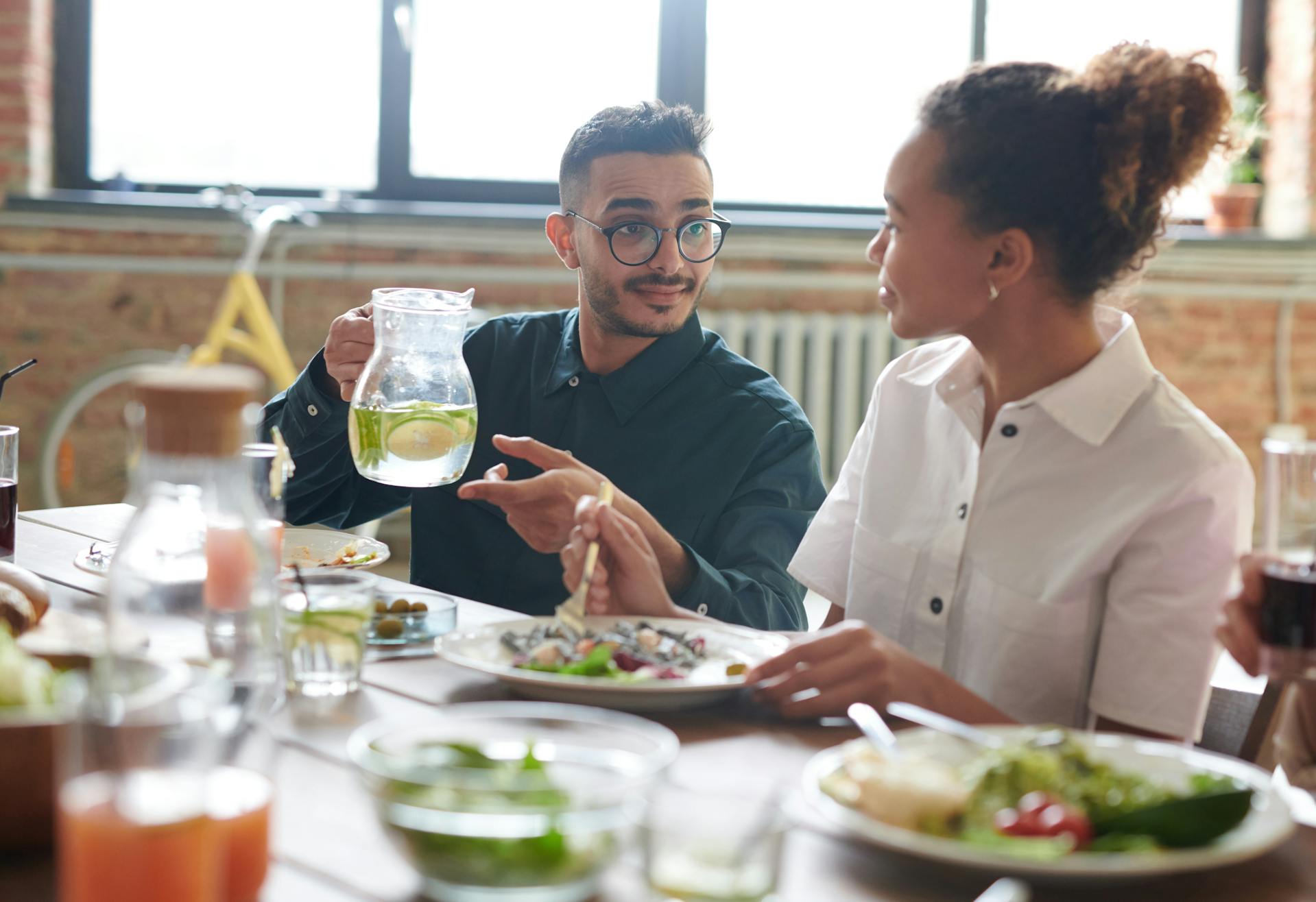 A man offers a jug of fresh water to a woman as they sit at a table together eating. A man offers a jug of fresh water to a woman as they sit at a table together eating.